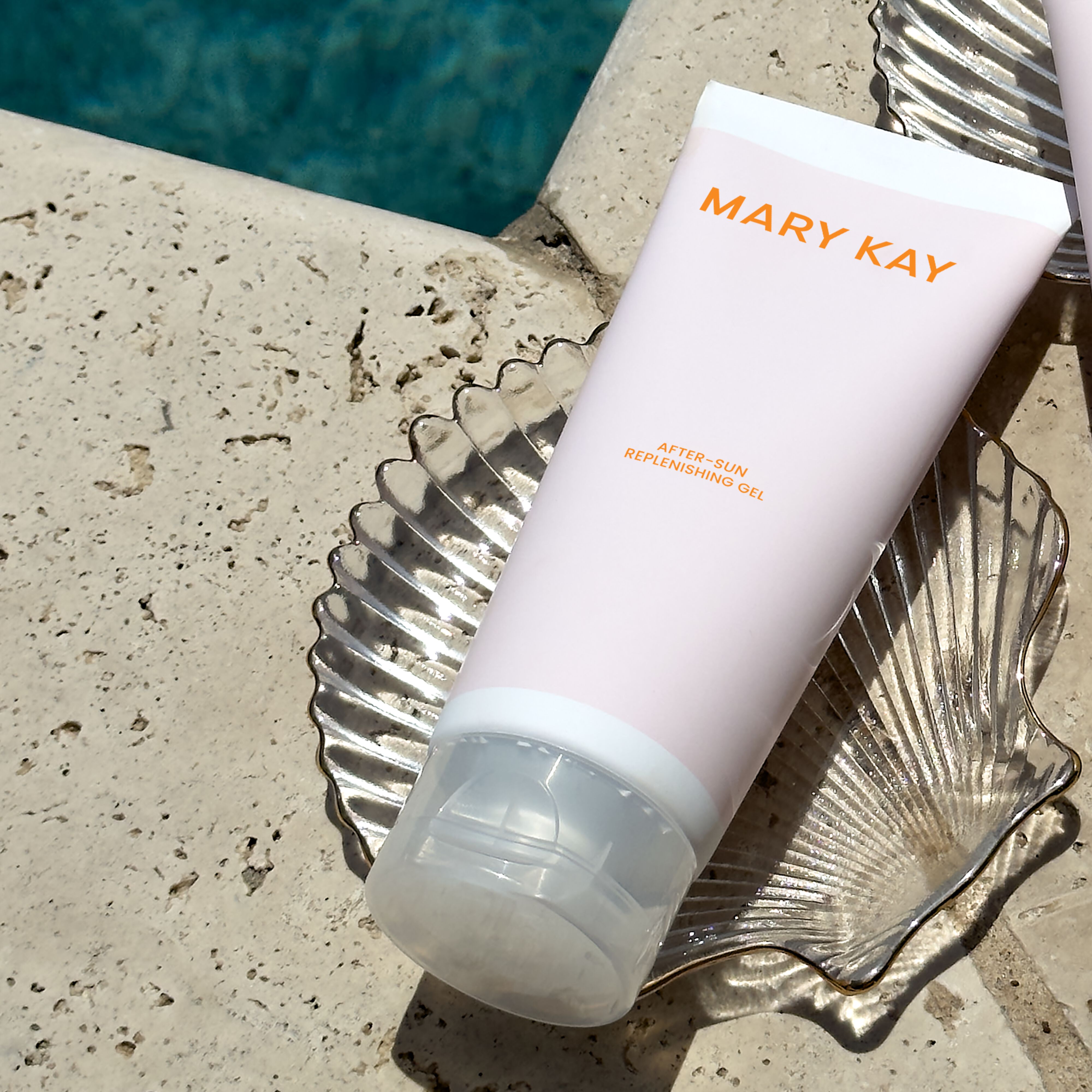 Mary Kay After-Sun Replenishing Gel tube resting on a textured stone pool edge beside a glass tray, with blue water visible in the background.